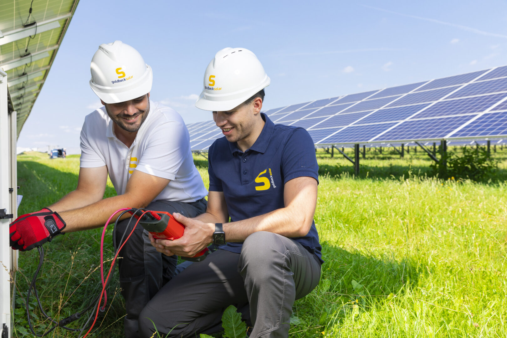 Zwei Mitarbeiter des O&M Teams von Goldbeck Solar beim Prüfen einer Solaranlage. Die Person links trägt ein weißes Shirt, die Person rechts ein blaues Shirt mit Goldbeck-Solar-Logo. Beide tragen Helme.,